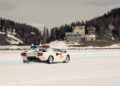 A white Lamborghini sports car with police lights drifts on a snowy track at I.C.E. St. Moritz in a mountainous area, with buildings and trees in the background.