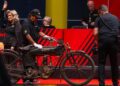 A vintage motorcycle is displayed at a Mecum Auctions event in Las Vegas. An auctioneer stands nearby, while a photographer captures the moment. Two people observe the captivating scene, highlighting the allure of this exclusive motorcycle auction.