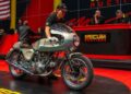 At a bustling Las Vegas setting, a person wheels a classic Ducati motorcycle on stage at the renowned Mecum Auctions, with attentive auctioneers and a striking red background enhancing the scene.
