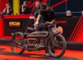 A person inspects an antique motorcycle at a Motorcycle Auction in Las Vegas, with auction staff in the background and a lively red and black theme.