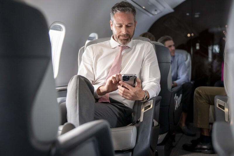 A man in a suit, immersed in his smartphone, enjoys the comforts of an airplane cabin. As he flies with Airshare aboard the luxurious Challenger 3500, he effortlessly blends business and leisure at 30,000 feet.