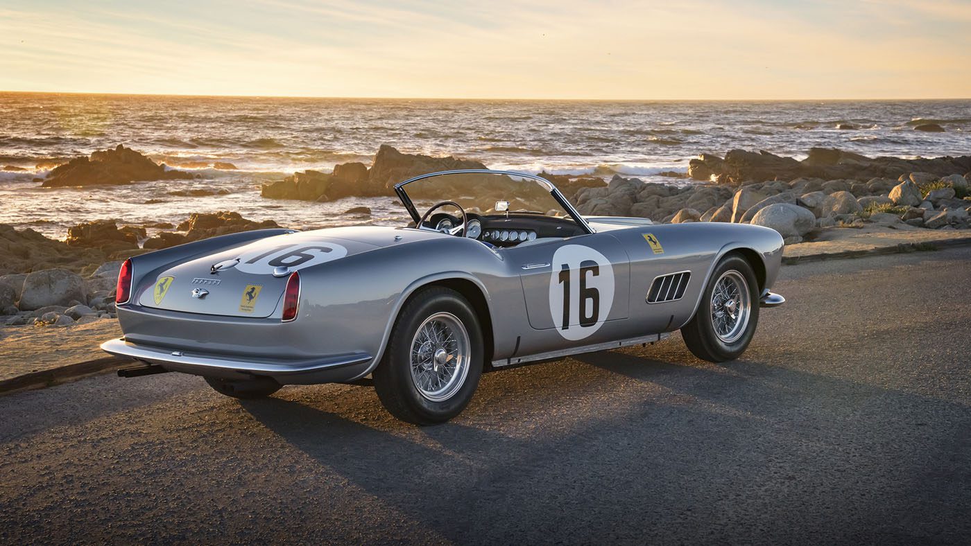 A vintage silver Ferrari 250 GT LWB California Spider, donned with racing number 16, is parked at the seashore at sunset. Its timeless allure complements the backdrop of sea and rocks, reminiscent of a scene straight from Amelia Island's prestigious Broad Arrow Auctions.