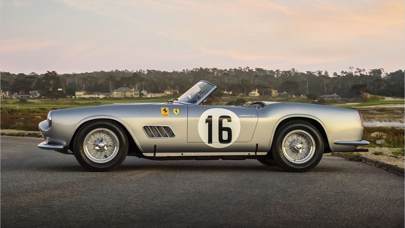 A vintage silver convertible sports car, reminiscent of a Ferrari 250 GT California Spider, marked with the number 16, is parked on a coastal road with houses and trees in the background. It looks like it’s ready to be showcased at an event like the Amelia Island Auctions.