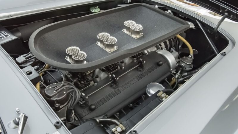 Close-up of a Ferrari 250 GT engine with three mesh-covered air filters on top. The engine, reminiscent of those seen at Amelia Island Auctions, is fitted neatly within the vehicle's open hood, showcasing various mechanical components and wiring.