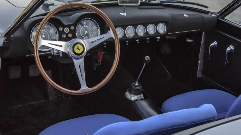 Interior of a vintage Ferrari 250 GT California Spider featuring a wooden steering wheel with a yellow emblem, classic round speedometers, manual gear stick, and blue seat covers. This timeless beauty would be a showstopper at any Amelia Island Auction.