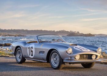 A vintage Ferrari sports car, reminiscent of the classic California Spider, proudly displays the number 16 as it rests by the ocean under a partly cloudy sky, with Amelia Island's rocky coastline painting a picturesque background.