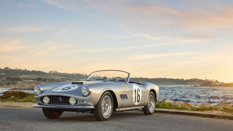 A silver classic sports car, reminiscent of a Ferrari 250 GT California Spider with the number 16 on the door, is parked by the seaside at sunset. This timeless beauty glows in the evening light, evoking memories of its glory days and perhaps a moment at an Amelia Island Auction.