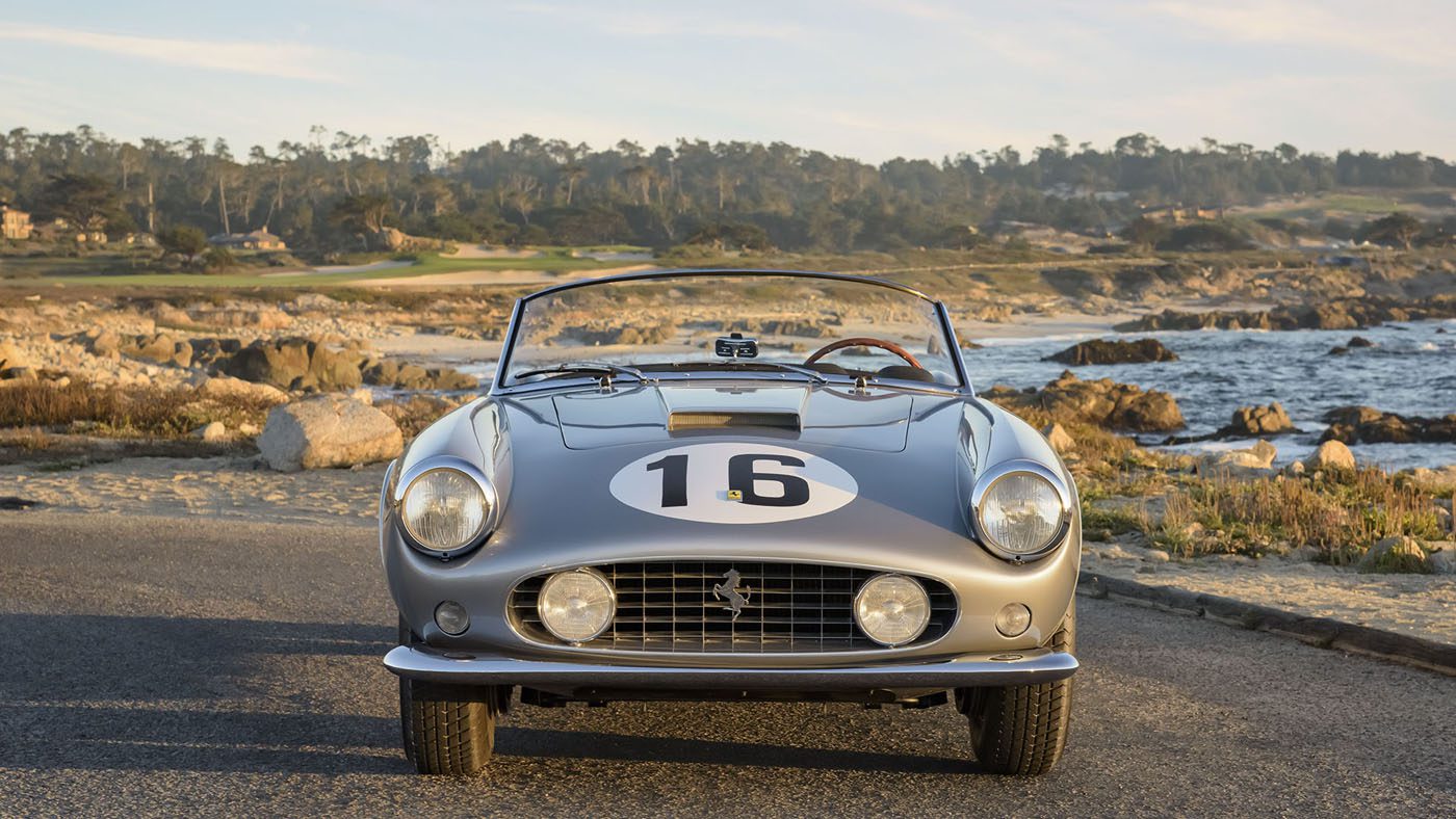 A vintage silver Ferrari 250 GT convertible car with the number 16 on the hood, parked along a coastal road with rocks and trees in the background, reminiscent of treasures found at prestigious Amelia Island Auctions.