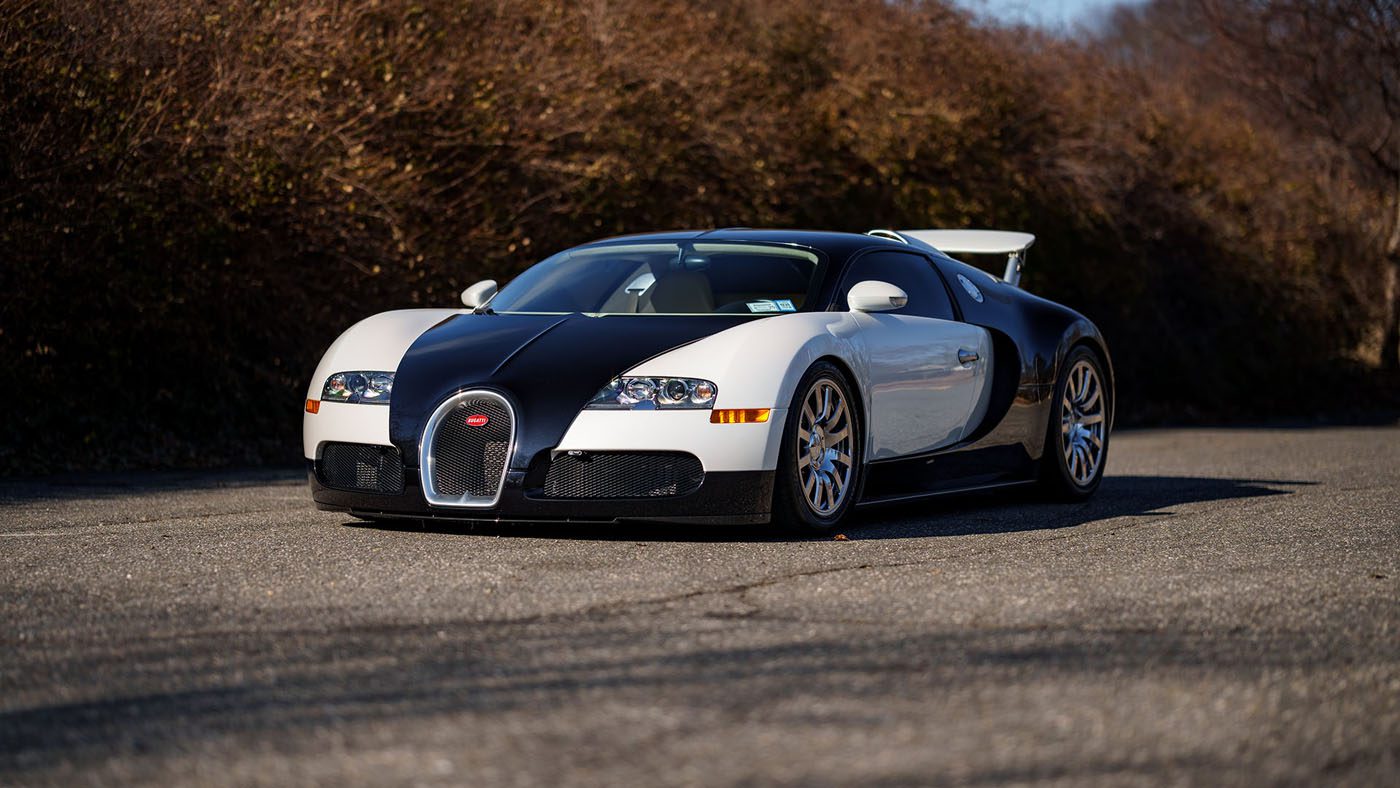 A sleek black and white sports car, resembling a Bugatti Veyron, is parked on a deserted road, surrounded by dry bushes and trees.