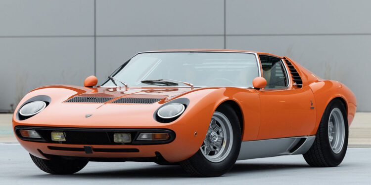 A 1971 Lamborghini Miura, with its orange vintage allure and sleek design, is poised at an angle on the gray surface. This classic beauty could easily attract attention at prestigious events like Broad Arrow Auctions or Amelia Island.