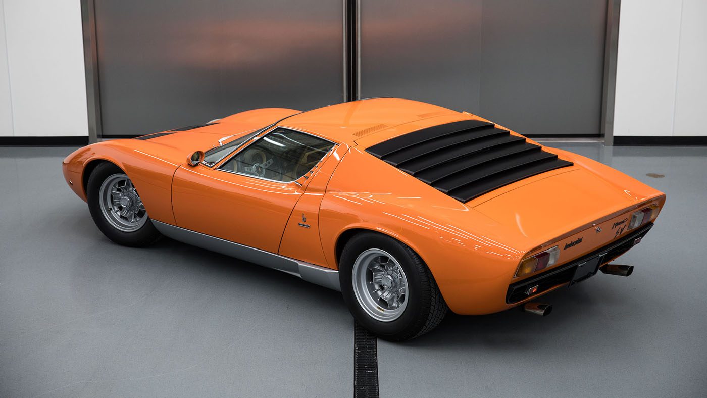 An orange classic sports car with a sleek design, reminiscent of the Lamborghini Miura, is viewed from the rear in an indoor setting. This beauty could easily be a star at Amelia Island or featured by Broad Arrow Auctions.