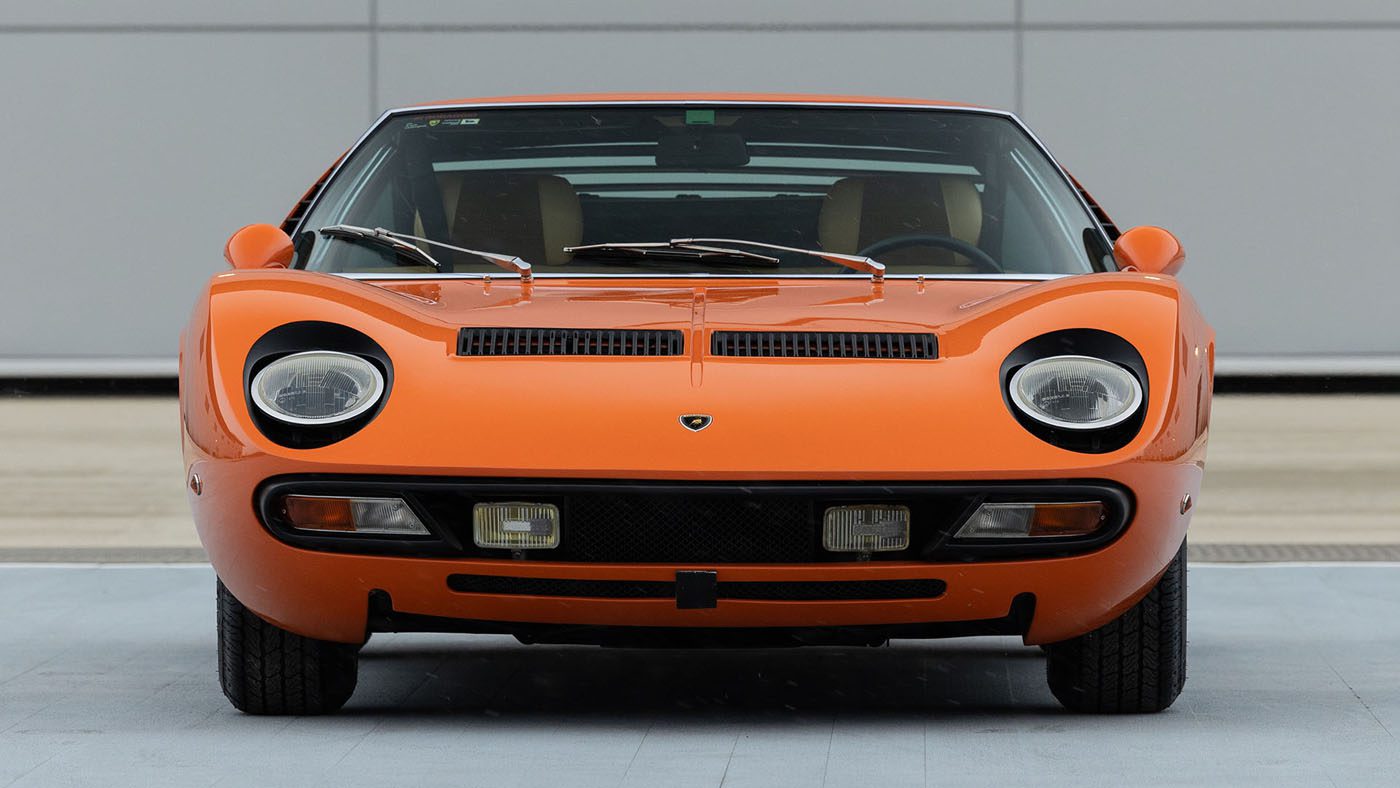 Front view of a Lamborghini Miura, an orange classic sports car with black bumper details and visible headlights, parked on a paved area in front of a plain wall. A sleek design that might catch eyes at an event like Amelia Island or Broad Arrow Auctions.