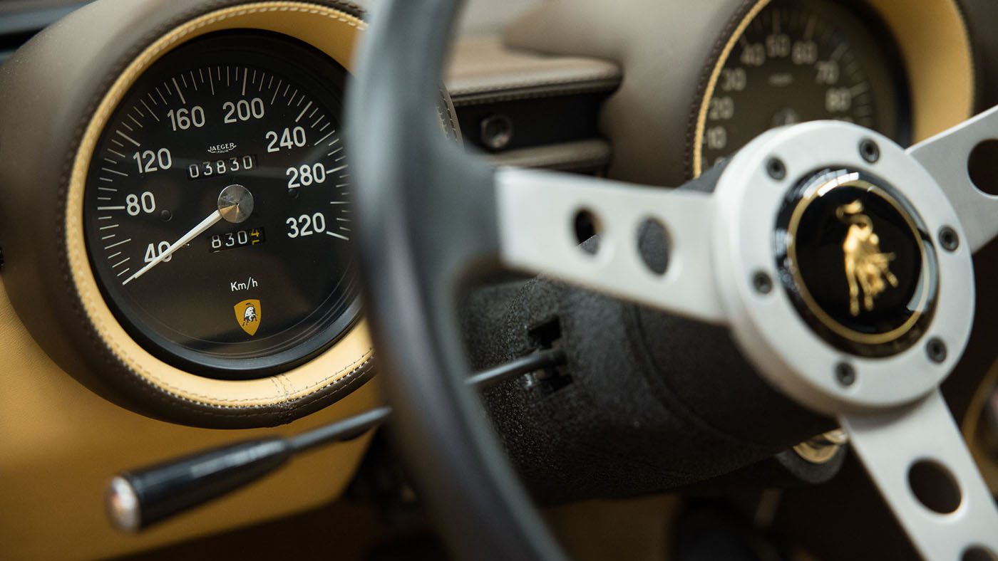 Close-up of a car dashboard reveals a speedometer maxing out at 280 km/h beside a circular steering wheel adorned with the logo of the iconic 1971 Lamborghini Miura, a classic celebrated for its sleek design often seen at Broad Arrow Auctions.