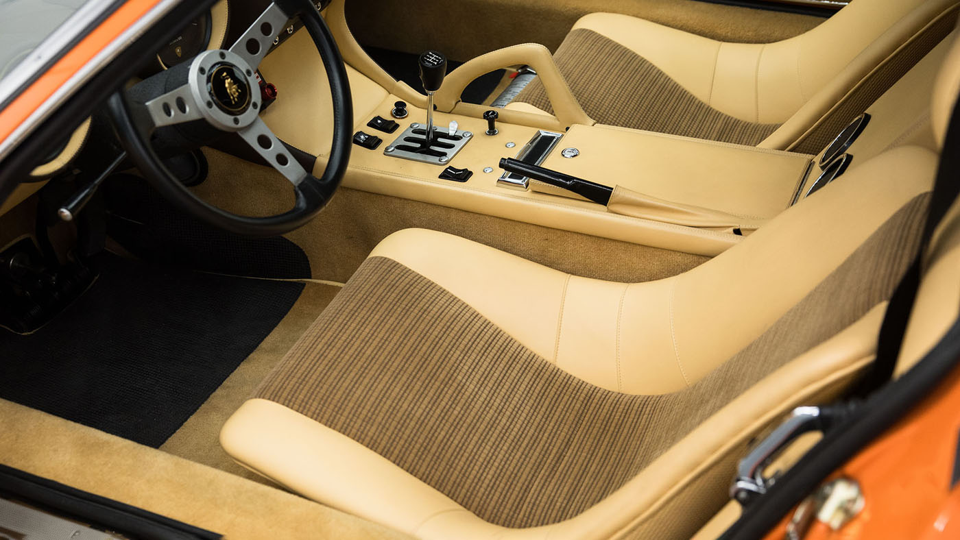 This vintage car interior evokes the classic elegance of a 1971 Lamborghini Miura P400 SV, showcasing tan striped upholstery, a black steering wheel, and manual gear shift. Picture this timeless beauty at an event like Broad Arrow Auctions on Amelia Island.