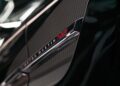 Close-up of the Vanquish Supercar's Aston Martin badge and V12 emblem on the side, capturing its metallic finish as a light sculpture highlights its sleek contours.
