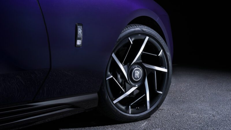 Close-up of a car's front wheel showing a black alloy rim with a Rolls-Royce logo in the center, set against a dark purple body.