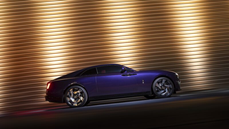 A sleek, dark purple luxury car drives past a lit-up, striped background at night.