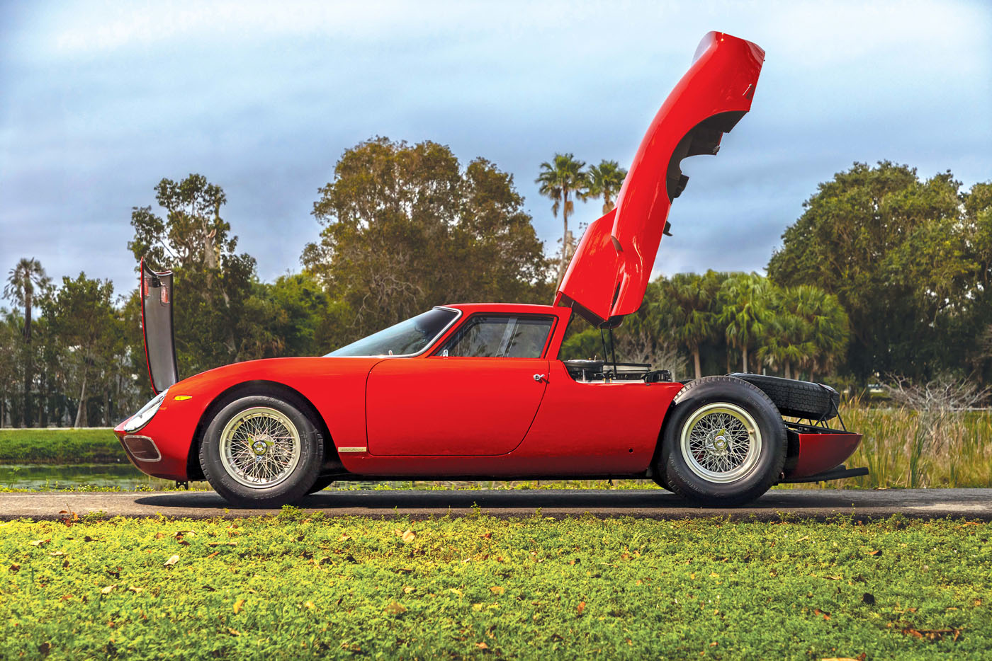 A red vintage Ferrari 250 LM, hailed as the best of the best, with its front hood open, is elegantly parked on a roadside, surrounded by trees—a scene worthy of the Cavallino Classic.