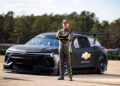 The race car driver stands confidently with arms crossed in front of a sleek black Chevrolet NASCAR Prototype on the track, surrounded by lush greenery.