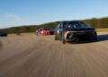 Race cars speed along a curved track, with a sleek black car in the foreground and a vibrant red Chevrolet Blazer EV following closely behind.
