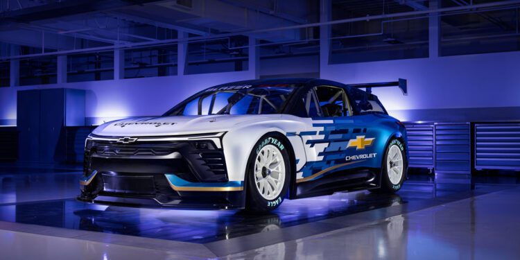 The Chevrolet race car, a stunning NASCAR Prototype in white and blue with graphic design, is displayed indoors on a reflective floor under overhead lights.