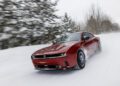 A red Dodge Charger, with its powerful AWD system, races through a snowy, forested road at high speed, leaving a blur of snow and trees in its wake.