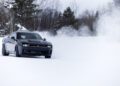 A black Dodge Charger, the world's only AWD muscle car, drifts across a snow-covered landscape with trees in the background, creating a stunning cloud of snow.