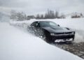 A black Dodge Charger, renowned as the world's only AWD muscle car, drives through a thick layer of snow on the road, snow spraying around it. In the background, snow-covered trees and buildings complete this wintry scene.