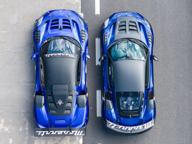 Two blue Maserati sports cars, viewed from above. One has a transparent engine cover. Both have large rear spoilers and visible rear air vents, parked side by side on a gray surface.