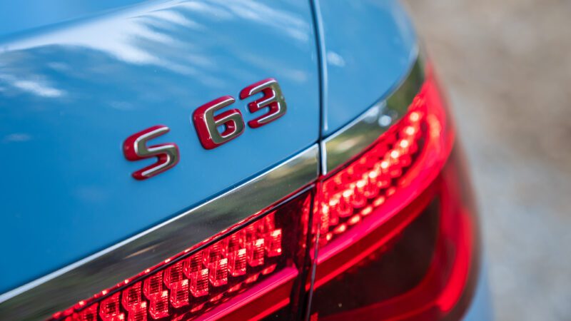 Close-up of the rear end of a car featuring the badge "S 63" and a lit taillight. The car is painted in a light blue color.