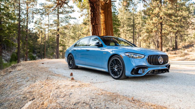 A blue luxury car is parked on a forest road, surrounded by tall pine trees under a clear sky.