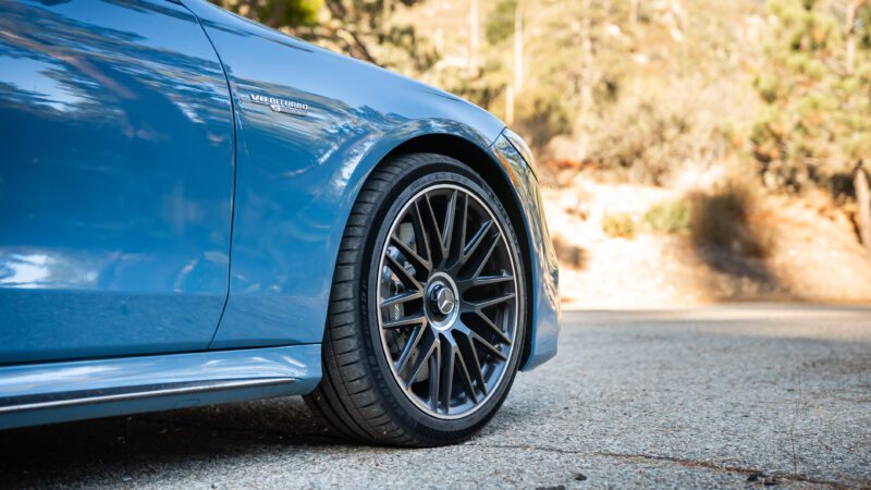 Close-up of a blue car's front left tire and rim, parked on a paved surface. The car features a "V8 BITURBO" emblem. Background shows blurred trees and a sunny setting.