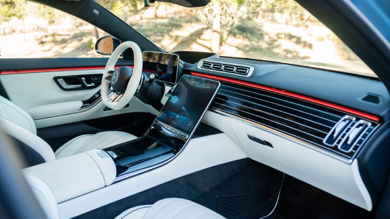 Modern car interior featuring a sleek dashboard, large touchscreens, and a steering wheel. The cabin has white leather seats and red accent lighting.