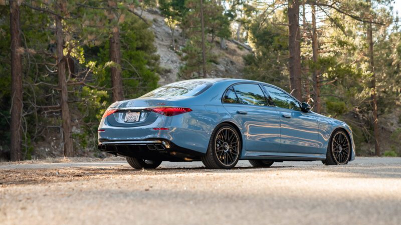 A blue Mercedes-AMG S63 sedan parked on a dirt road in a wooded area, viewed from the rear side under daylight.