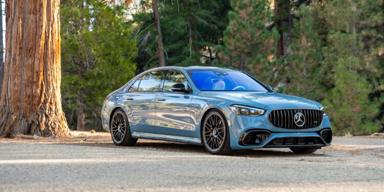 A blue luxury sedan parked on a forest road with tall trees in the background.