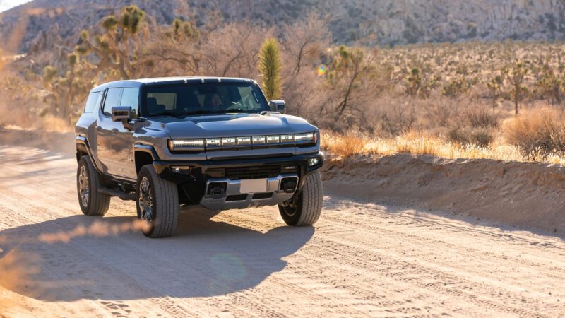 A black SUV drives on a dusty, unpaved road in a desert landscape with sparse vegetation and distant rocky hills.