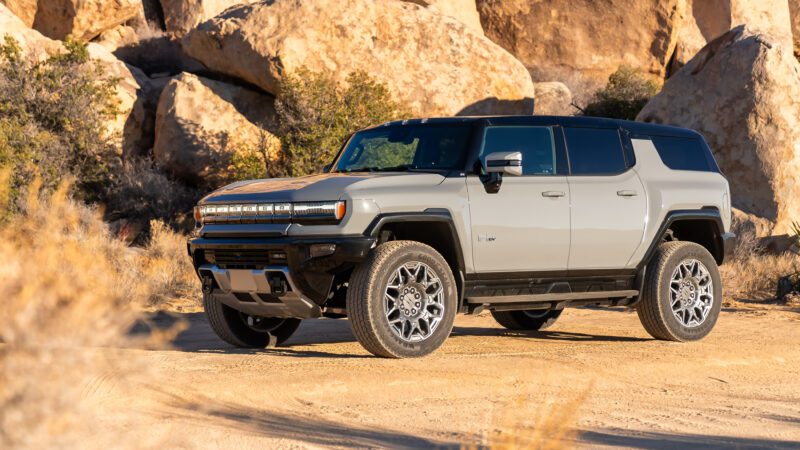 A gray SUV is parked on a desert terrain with large boulders in the background, under clear skies.