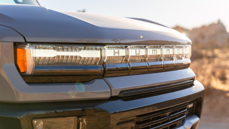 Close-up of the front grille of a Hummer vehicle, featuring the brand name in bold letters, with a blurred outdoor background and sunlight reflecting on the surface.