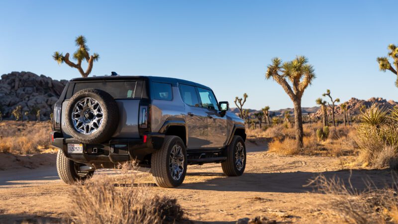 An off-road vehicle parked on a desert trail with rocky terrain and Joshua trees in the background under a clear blue sky.