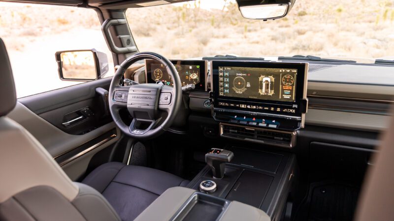 Interior view of a vehicle cabin featuring a digital dashboard with multiple screens, a steering wheel, and a mix of black and beige upholstery. Desert landscape visible through the windows.