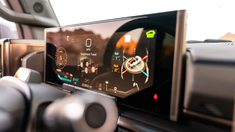 Close-up of a vehicle's digital dashboard displaying speed, suspension travel, and various indicators. The screen is glossy with reflections.