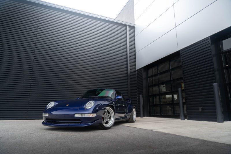 A dark blue Porsche 993 sports car is parked on a concrete surface next to a black metal-paneled building, showcasing its sleek design. This remarkable 911 GT2 Tribute is now for sale, offering a rare opportunity to own an iconic piece of automotive history.