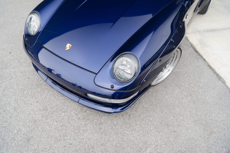 Close-up mobil sport Porsche 911 GT2 biru, menampilkan kap depan, lampu depan, dan bagian dari fender pada permukaan beton abu-abu dan putih.