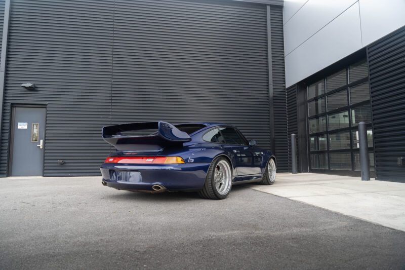 A sleek Porsche 993 with the 911 GT2 Tribute package, featuring a large rear spoiler, is parked near a modern building with dark corrugated panels and glass doors. It's an enticing scene for car enthusiasts, especially now that this beauty is for sale.