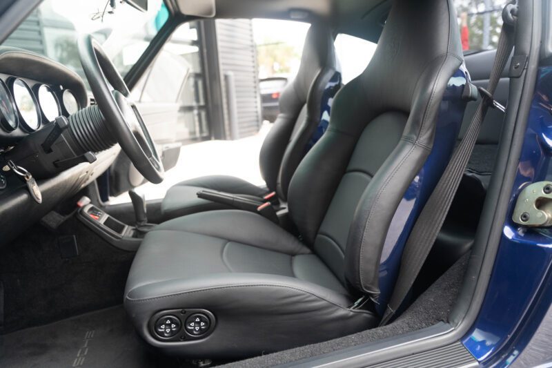 The blue interior of this Porsche 993 features sleek black leather seats, a stylish steering wheel, and a modern dashboard. The open door reveals seat adjustment controls and car keys in the ignition—a classic tribute for sale that combines elegance with performance.