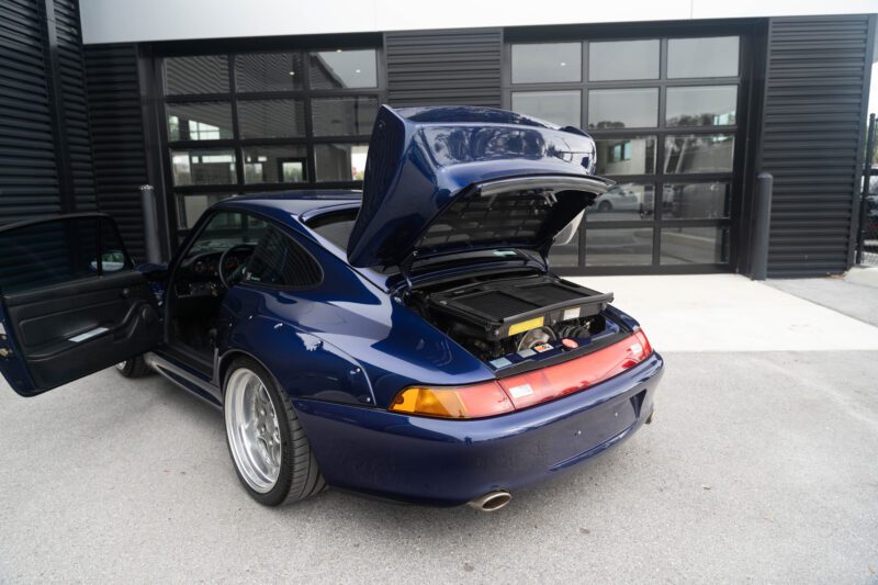 A blue Porsche 993 with its engine compartment open sits elegantly outside a modern building, effortlessly capturing attention.