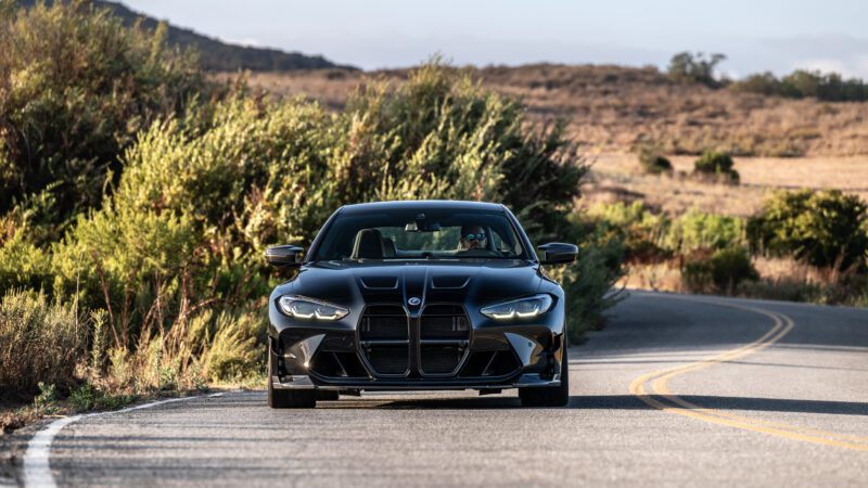 A black sports car drives on a winding rural road surrounded by greenery, under a clear sky.