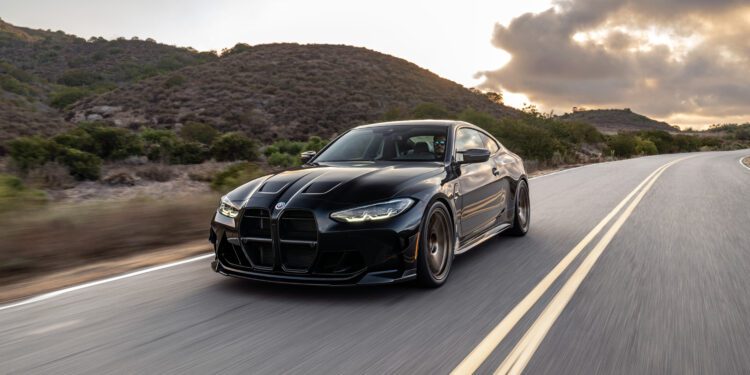 A black sports car drives on a scenic, empty road surrounded by hills and a cloudy sky.