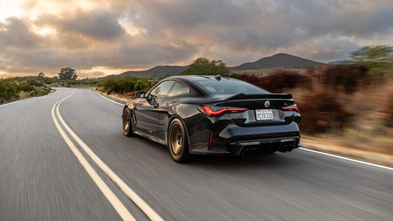 A black sports car drives on a winding road with mountains and cloudy sky in the background.