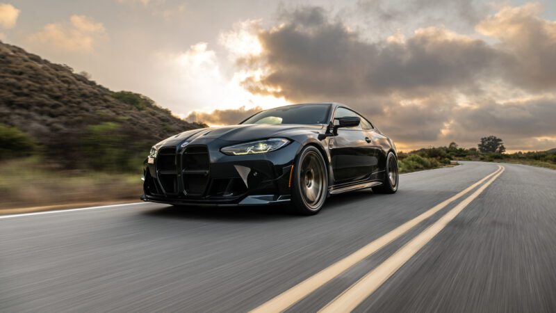 A black sports car drives on a winding road with hills and clouds in the background.